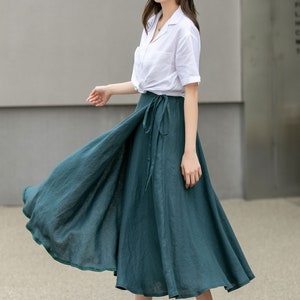 May include: A woman wearing a white button-down shirt, a wide-brimmed brown hat, and a teal green wrap skirt. She is standing on a sidewalk in front of a building.