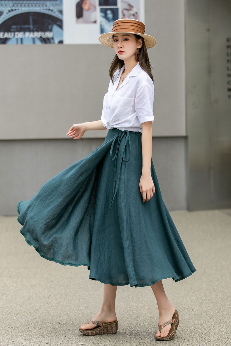 May include: A woman wearing a white button-down shirt, a wide-brimmed brown hat, and a teal green wrap skirt. She is standing on a sidewalk in front of a building.