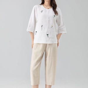 May include: A white linen top and beige linen capri pants. The top has a round neck, elbow-length sleeves, and embroidered floral details. The capri pants are a neutral beige color. The outfit is casual and comfortable.