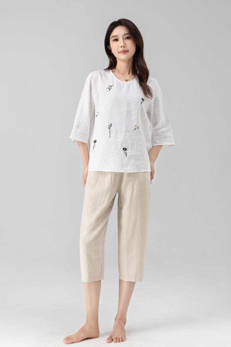 May include: A white linen top and beige linen capri pants. The top has a round neck, elbow-length sleeves, and embroidered floral details. The capri pants are a neutral beige color. The outfit is casual and comfortable.