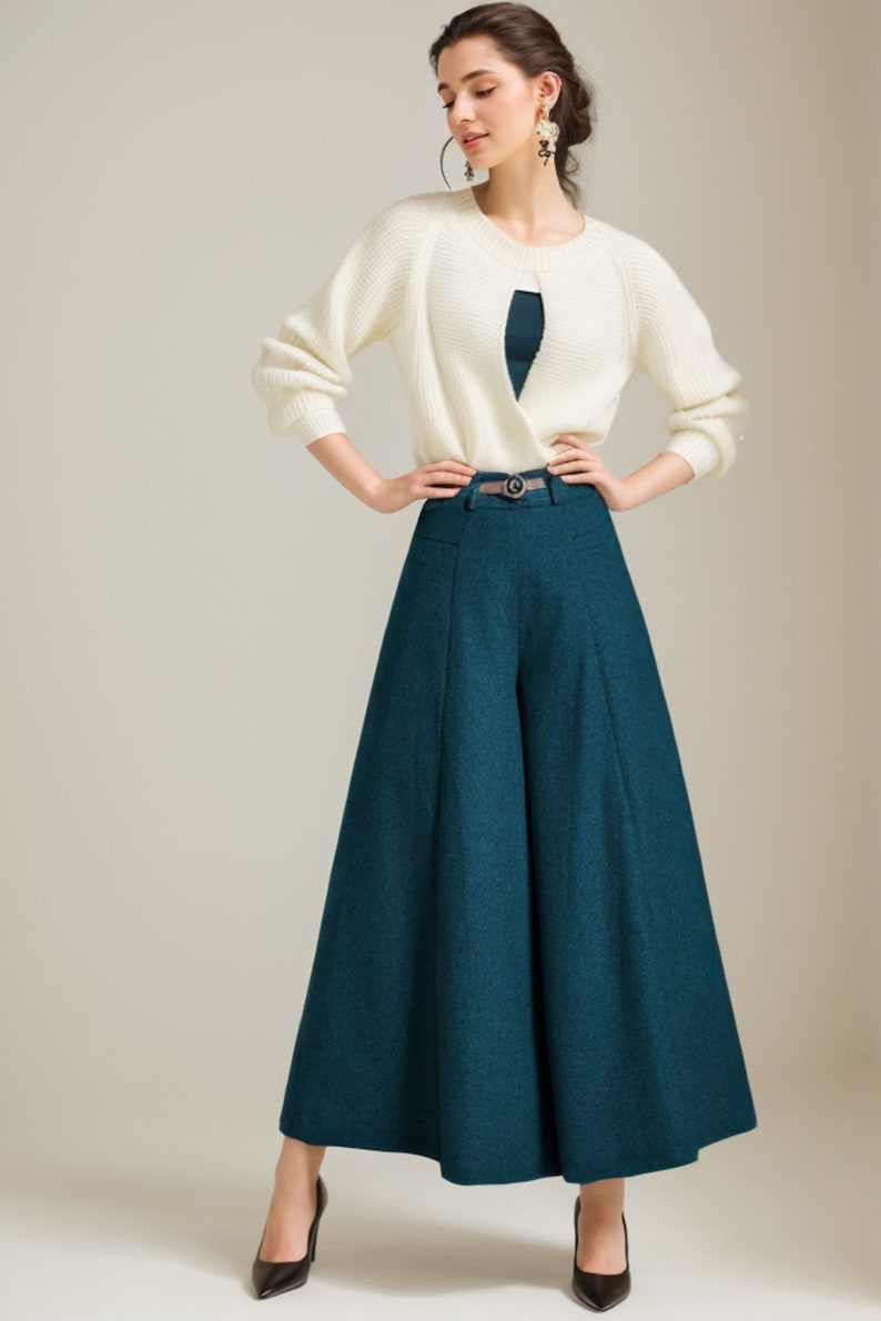 May include: A woman wearing a white knit sweater and teal wide-leg pants with a belt. She is standing on a light beige background.