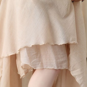 May include: Close-up of a beige, layered fabric garment. The fabric has a slightly textured appearance and a scalloped edge on the lower layers. The garment appears to be made of a lightweight material, possibly linen or a similar fabric.