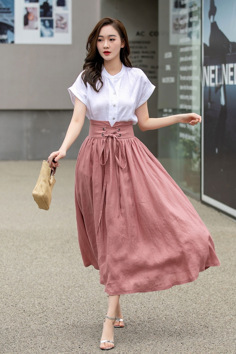 May include: A woman wearing a white button-down shirt with a pink skirt tied with a bow at the waist. She is holding a straw bag and walking on a sidewalk.
