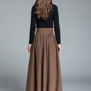 May include: A brown high-waisted skirt with a pleated design. The skirt is made of a thick, textured fabric and falls to the ankles. The skirt is paired with black heeled boots.