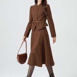 May include: A brown skirt suit with a fitted jacket and a-line skirt. The jacket has a high collar, gold buttons, and a matching belt. The skirt is knee-length and has a slight flare.