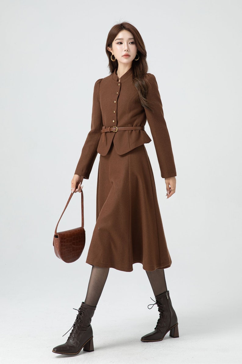 May include: A brown skirt suit with a fitted jacket and a-line skirt. The jacket has a high collar, gold buttons, and a matching belt. The skirt is knee-length and has a slight flare.