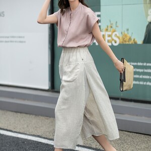 May include: A woman wearing a pink short-sleeved blouse, a wide-legged linen skirt, a brown hat, and brown wedge sandals. She is carrying a straw bag.
