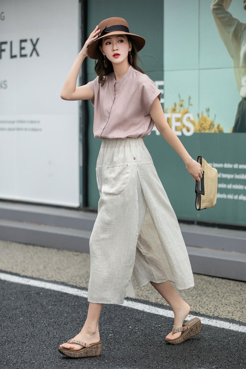May include: A woman wearing a pink short-sleeved blouse, a wide-legged linen skirt, a brown hat, and brown wedge sandals. She is carrying a straw bag.
