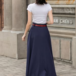 May include: A woman wearing a white t-shirt, navy blue maxi skirt, and a red belt. She is also wearing a black hat with a white band. The outfit is completed with clear heeled sandals. The background features a brick walkway and a building.