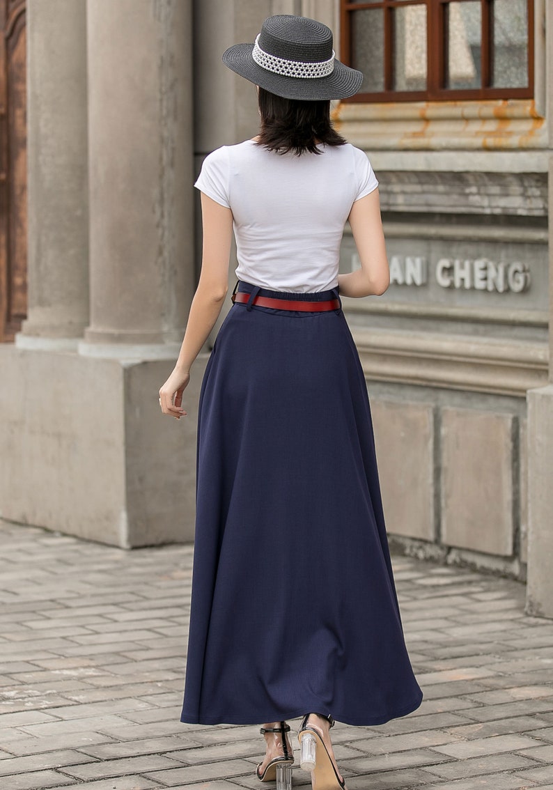 May include: A woman wearing a white t-shirt, navy blue maxi skirt, and a red belt. She is also wearing a black hat with a white band. The outfit is completed with clear heeled sandals. The background features a brick walkway and a building.
