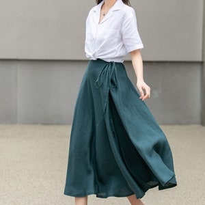 May include: A woman wearing a white linen shirt with a knotted front, a dark green wrap skirt, and a brown straw hat. She is wearing brown sandals with a woven design.