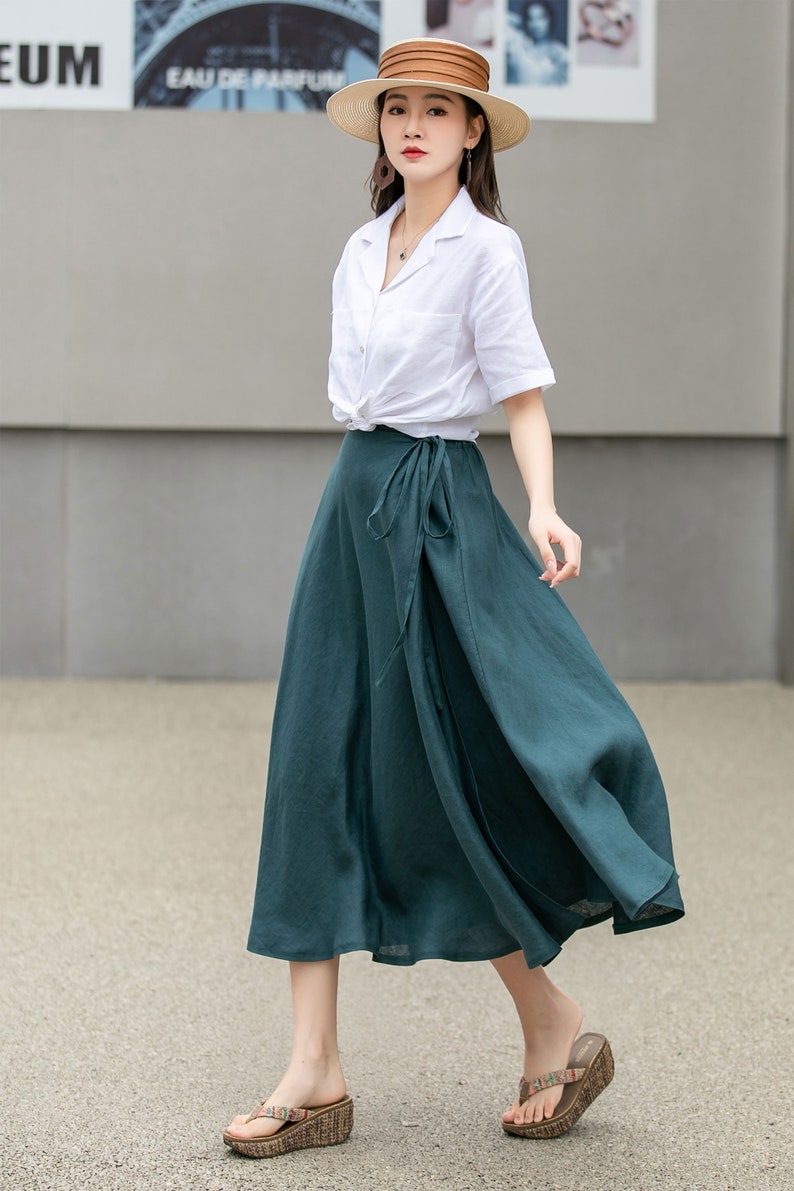 May include: A woman wearing a white linen shirt with a knotted front, a dark green wrap skirt, and a brown straw hat. She is wearing brown sandals with a woven design.