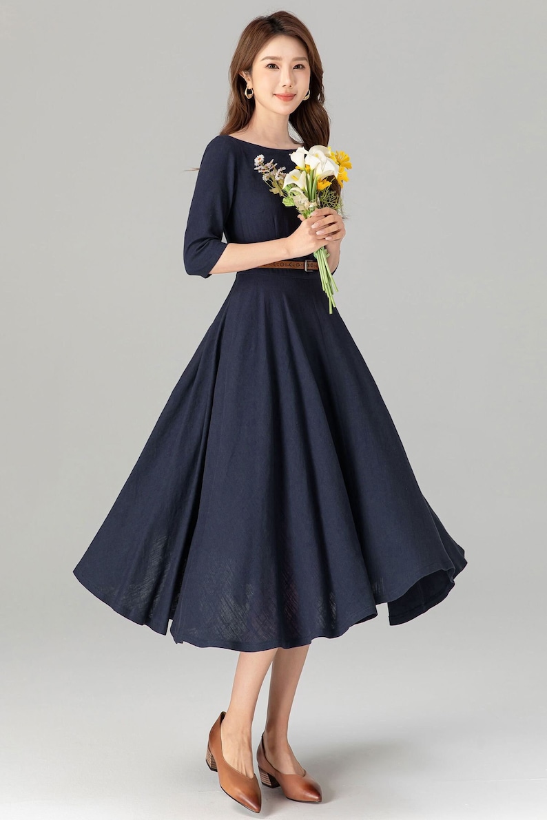 May include: A navy blue midi dress with a floral embroidery detail on the neckline. The dress has a fitted bodice and a full skirt. It features a brown belt at the waist. The model is wearing brown heeled shoes.