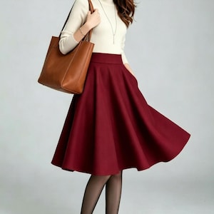 May include: A cream-colored long-sleeve mock neck top paired with a high-waisted, knee-length burgundy flared skirt. The outfit includes a brown leather tote bag, sheer black tights, and dark-colored high-heeled pumps with a thin silver necklace.