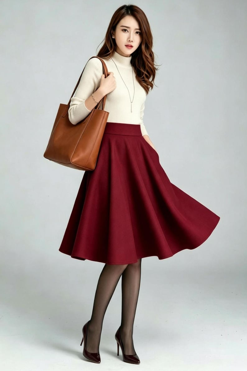 May include: A cream-colored long-sleeve mock neck top paired with a high-waisted, knee-length burgundy flared skirt. The outfit includes a brown leather tote bag, sheer black tights, and dark-colored high-heeled pumps with a thin silver necklace.