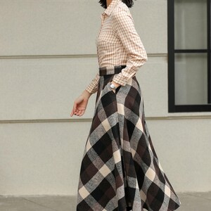 May include: A woman wearing a tan plaid button-down shirt and a long, plaid skirt in brown, black, and cream. She is also wearing brown boots with laces. The outfit is suitable for fall or winter.