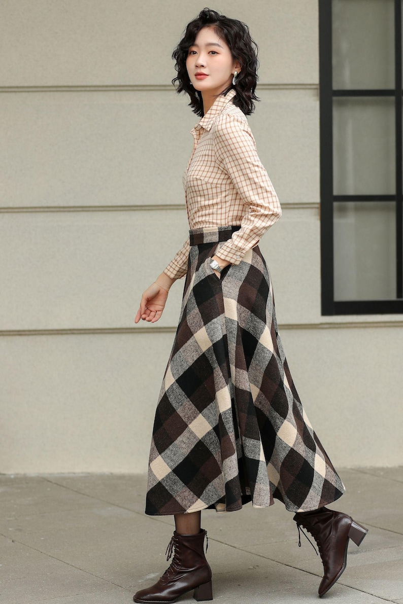 May include: A woman wearing a tan plaid button-down shirt and a long, plaid skirt in brown, black, and cream. She is also wearing brown boots with laces. The outfit is suitable for fall or winter.