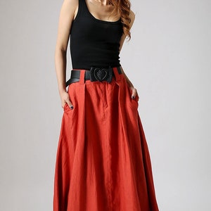 May include: A red maxi skirt with a black belt featuring a heart-shaped buckle. The skirt has a pleated design and pockets on the sides.
