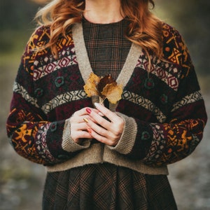 Pu&ograve; includere: Una donna indossa un cardigan a motivi marroni, rossi e gialli con un vestito a quadri. Tiene in mano un piccolo mazzo di foglie secche.