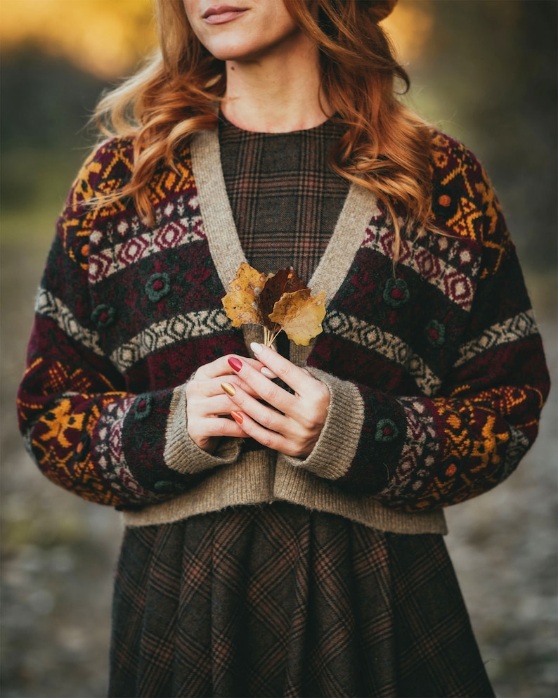 Pu&ograve; includere: Una donna indossa un cardigan a motivi marroni, rossi e gialli con un vestito a quadri. Tiene in mano un piccolo mazzo di foglie secche.