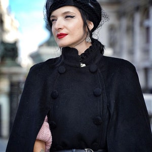 May include: A woman wearing a black cape with a black velvet hat and a black belt with a silver D buckle. She is wearing a pink sweater under the cape.