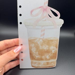 May include: A watercolor-style illustration of an iced coffee drink in a clear plastic cup, with a pink bow and a striped straw. The image is printed on a sheet of paper with three holes for a binder. The drink has ice cubes and a light brown color.