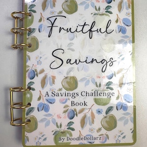 May include: A savings challenge book titled Fruitful Savings by DoodleDollarz with a white cover featuring a pattern of green apples and blue plums. The book is bound with gold-colored metal rings on the left side and has a green border.