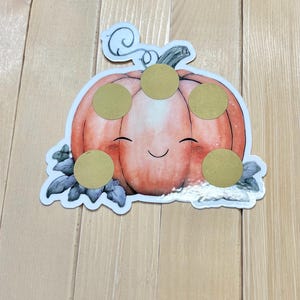 Op de afbeelding: Een sticker met een lachende cartoon pompoen met een steel en bladeren. De pompoen is oranje met een lichtroze midden en heeft vijf gouden cirkelstickers op zijn gezicht. De sticker ligt op een licht houten oppervlak.
