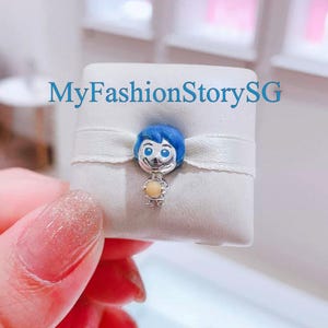 May include: A silver charm depicting a cartoon character with blue hair and a yellow orb. The charm is attached to a white ribbon on a white square. The text "MyFashionStorySG" is visible.