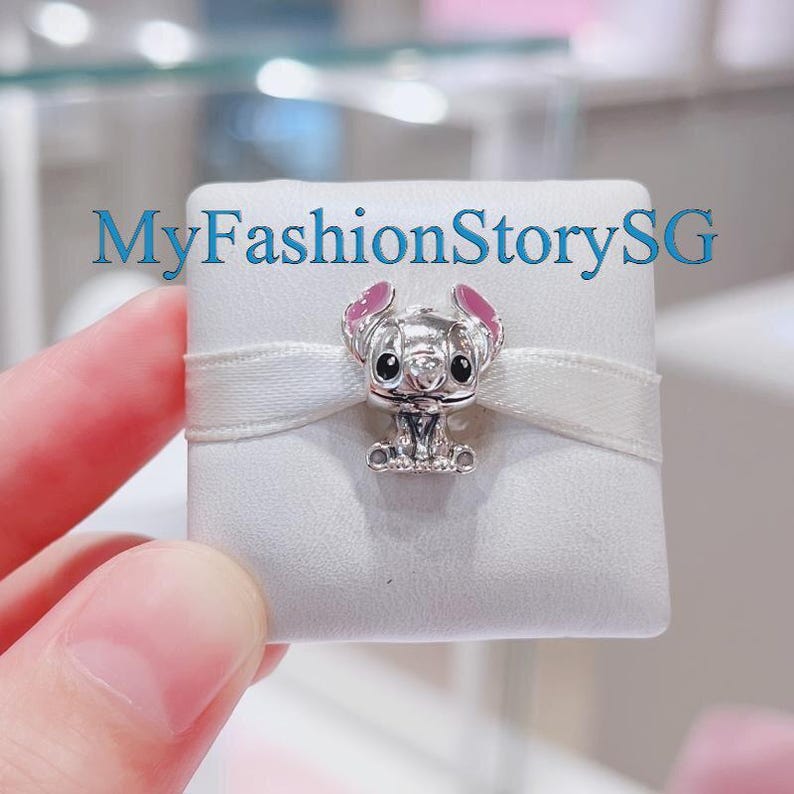 Pu&ograve; includere: Un ciondolo d'argento con Stitch del film Disney Lilo & Stitch. Stitch &egrave; seduto con le braccia distese e ha le orecchie rosa. Il ciondolo &egrave; in una scatola bianca con un nastro bianco.