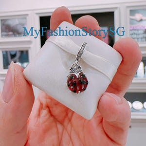 Puede incluir: Un charm de plata en forma de mariquita con esmalte rojo y detalles de cristal transparente. El charm está sobre un fondo blanco.