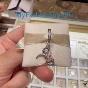 May include: A silver charm featuring a Mickey Mouse head design, with a pink and white bow adorned with clear crystals. The charm is displayed on a white background.