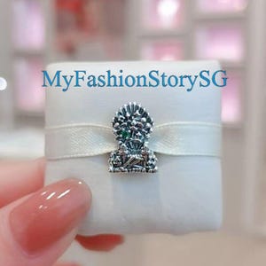 Puede incluir: Un charm de plata con un diseño floral detallado y una pequeña gema verde. El charm está sobre una caja blanca con una cinta blanca. El texto "MyFashionStorySG" es visible.