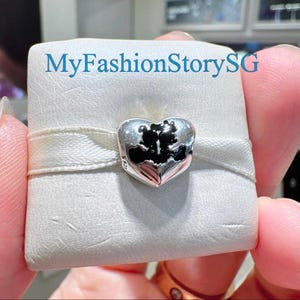 May include: A silver heart-shaped charm with a black silhouette design. The charm is held on a white satin ribbon. The text "MyFashionStorySG" is visible in the background.