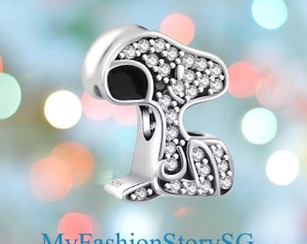 May include: A silver charm in the shape of Snoopy, the cartoon dog, with a white hat and black ears. The charm is encrusted with small, clear crystals. The text "MyFashionStorySG" is at the bottom.