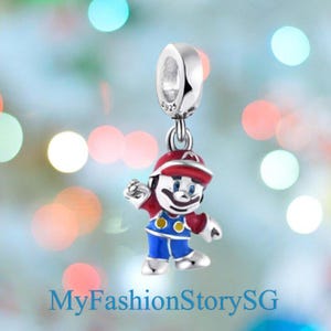 Puede incluir: Un charm de plata con un personaje de dibujos animados con gorro rojo y peto azul. El charm está suspendido de un lazo plateado. El texto "MyFashionStorySG" está en la parte inferior de la imagen.