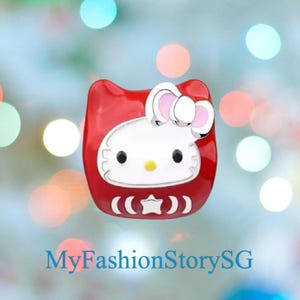 May include: A red and white Hello Kitty-themed accessory with a bow, black eyes, and a yellow nose. The accessory has a rounded shape with a star and bracket design at the bottom. The text "MyFashionStorySG" is at the bottom.