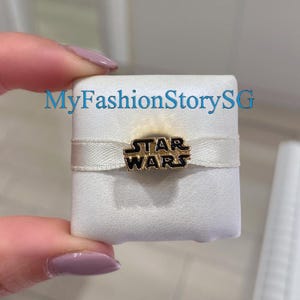 May include: A gold-toned metal charm featuring the black-filled Star Wars logo text, centred on a horizontal white ribbon band. The charm is mounted on a square white display cushion. The text MyFashionStorySG is visible in blue.