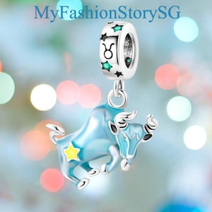 May include: A silver charm with a light blue bull pendant, a yellow star, and a ring with green stars and the Taurus symbol. The charm is suspended from a ring. The text "MyFashionStorySG" is at the top.