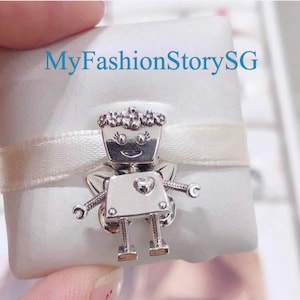 Puede incluir: Un charm plateado con forma de robot con corona, corazón y alas, presentado sobre una cinta de raso blanca y un cuadrado blanco. El texto "MyFashionStorySG" es visible en el fondo.