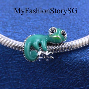 May include: A green chameleon charm with black eyes and silver accents. The charm is attached to a silver bracelet. The text "MyFashionStorySG" is at the top of the image. The charm is a vibrant accessory.