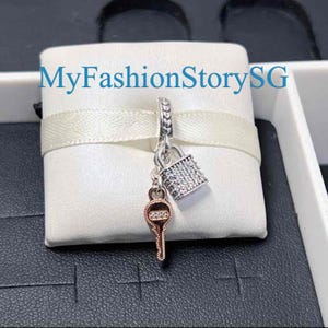 May include: A silver-toned charm featuring a padlock and a rose gold-toned key, both embellished with small clear stones. The charm hangs from a silver bail on a white ribbon. The text MyFashionStorySG is visible in blue across the top.