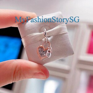 May include: A heart-shaped charm with a lock design. The charm is two-toned, with a rose gold frame and a silver-colored heart filled with small, sparkling stones. The charm is suspended from a silver-colored loop. The text "MyFashionStorySG" is visible.