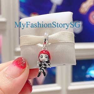 Puede incluir: Un charm de plata con un personaje de pelo rojo con un traje negro y una máscara plateada. El charm está colgado de una cinta blanca y se muestra sobre un fondo blanco.