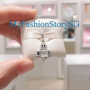 Puede incluir: Un charm de robot plateado con ojos negros y una cadena plateada. El charm está sobre un fondo blanco.