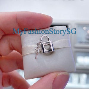 May include: A silver-colored charm featuring a padlock with a heart-shaped cutout and a small key. The charm is attached to a white square with a ribbon. The text "MyFashionStorySG" is visible in the background.