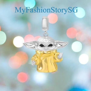 Puede incluir: Un colgante de plata y oro con un personaje de orejas grandes y una túnica amarilla. El dije tiene un lazo en la parte superior para sujetarlo a una pulsera o collar. El texto "MyFashionStorySG" está en el fondo.