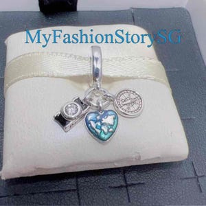 May include: Silver charm pendant with a camera, compass, and heart-shaped globe design. The camera has a clear gem, and the heart has blue and green accents. The text "MyFashionStorySG" is visible in the background.