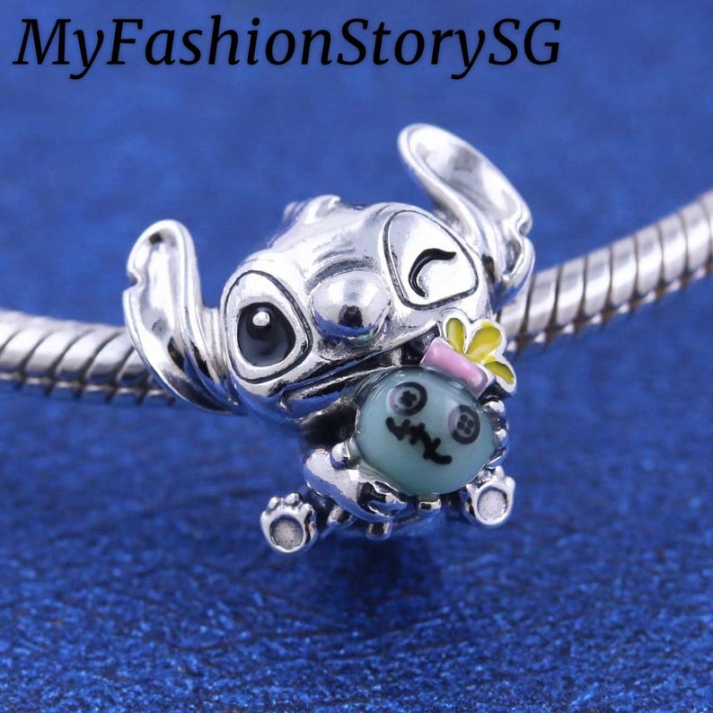 May include: A silver charm featuring a cartoon character holding a teal-colored object with a face. The charm has a flower and pink bow detail. The text "MyFashionStorySG" is in the top left corner.