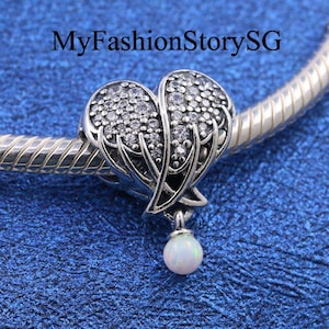 Puede incluir: Un charm de plata en forma de corazón con alas, adornado con piedras de circonita cúbica transparentes. Una pequeña opala blanca cuelga de la parte inferior del charm.